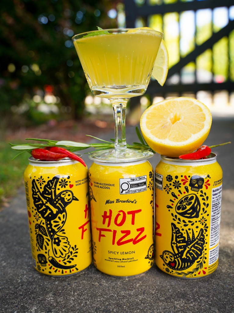 Three HOT FIZZ Spicy Lemon cans with a poured mocktail, lemon, chili, and herbs