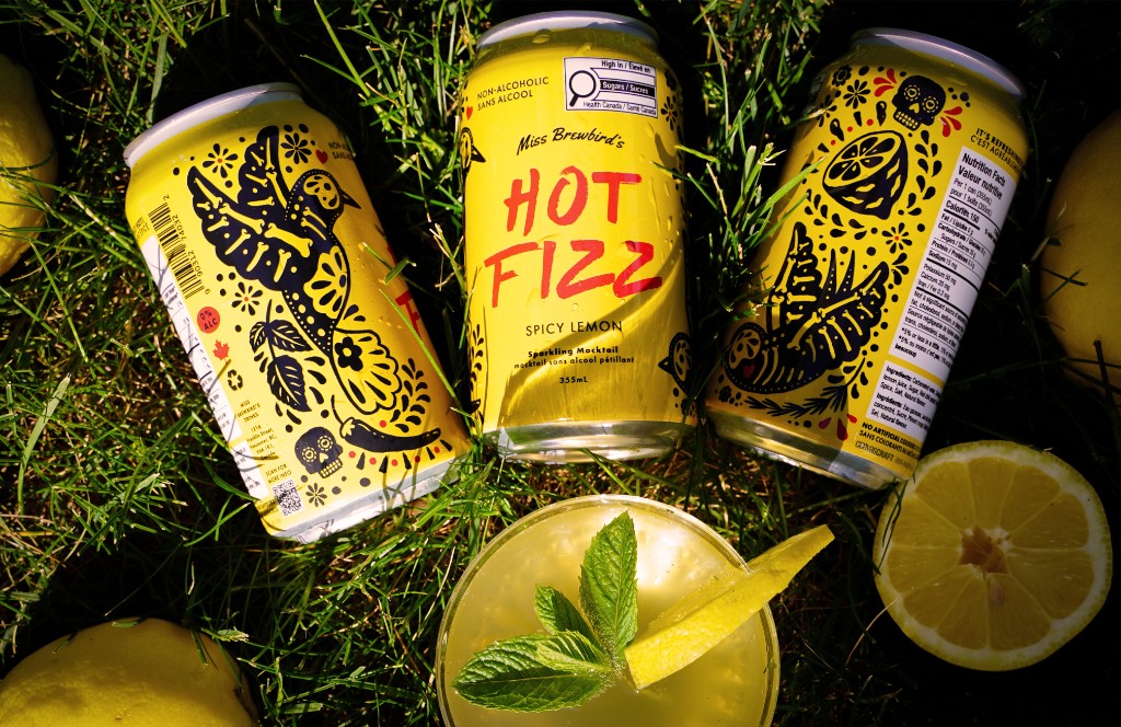 Three HOT FIZZ Spicy Lemon cans on grass with a poured drink and lemons